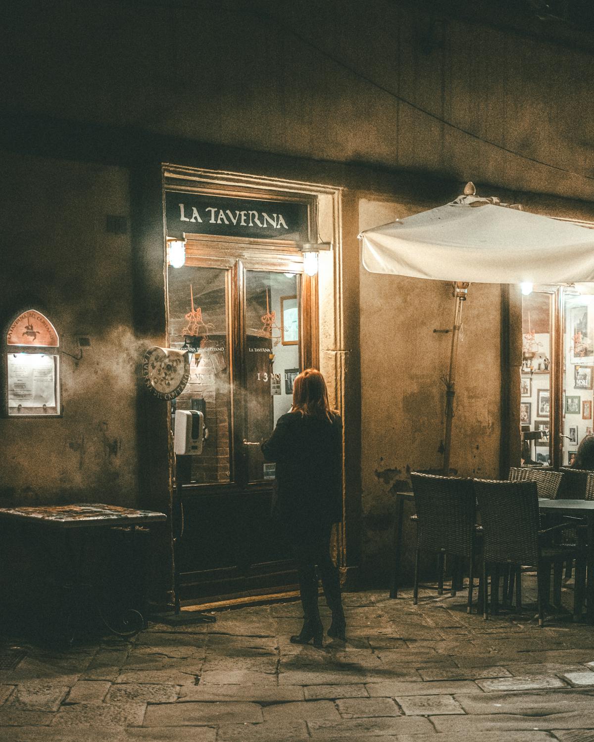 Person standing outside a warmly lit European taverna on a cobblestone street at night