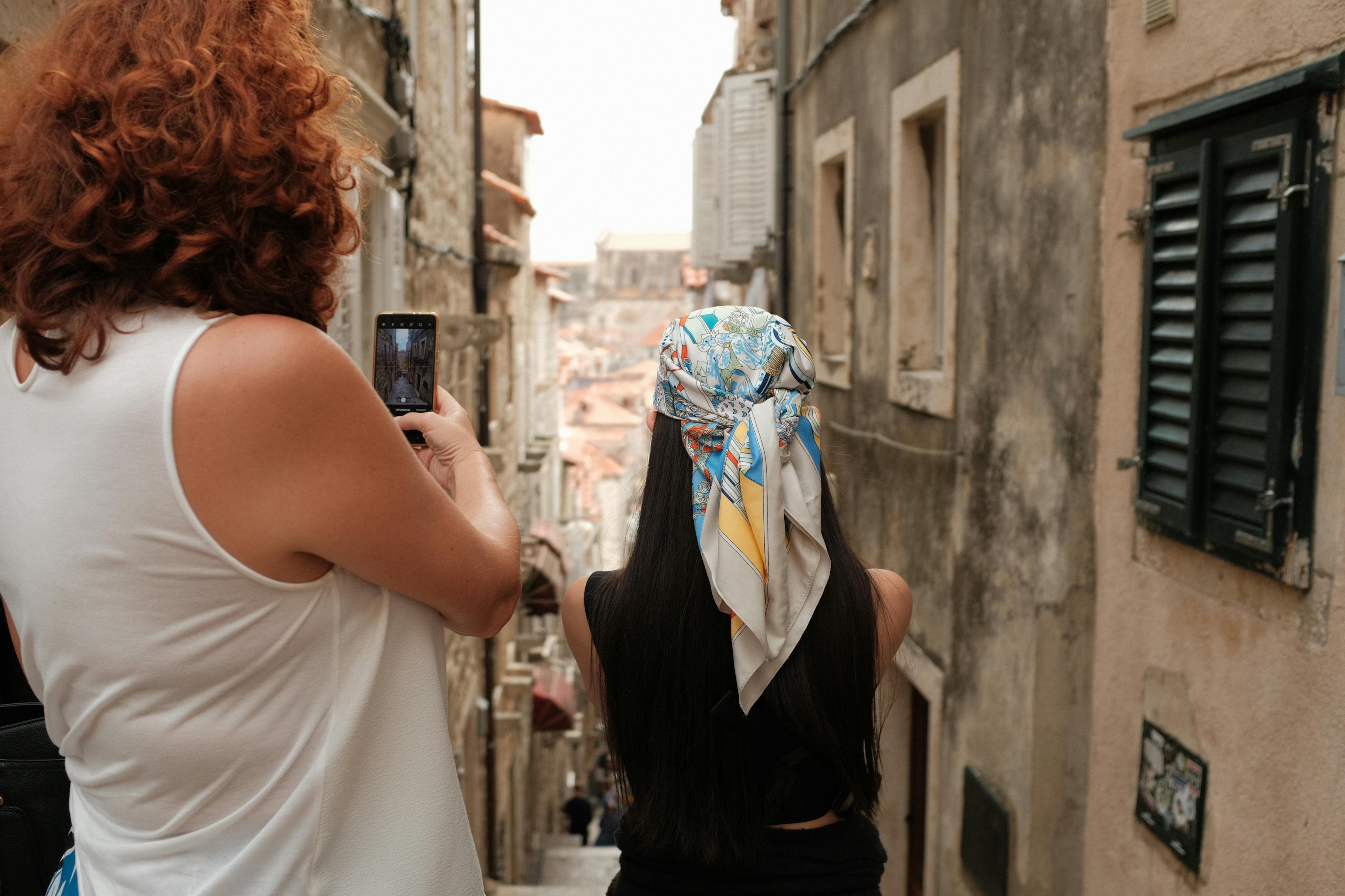 Two women explore a narrow alley in Dubrovnik's historic old town, capturing the moment on a smartphone