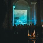 People silhouetted against an illuminated ocean art projection in a dark exhibition space