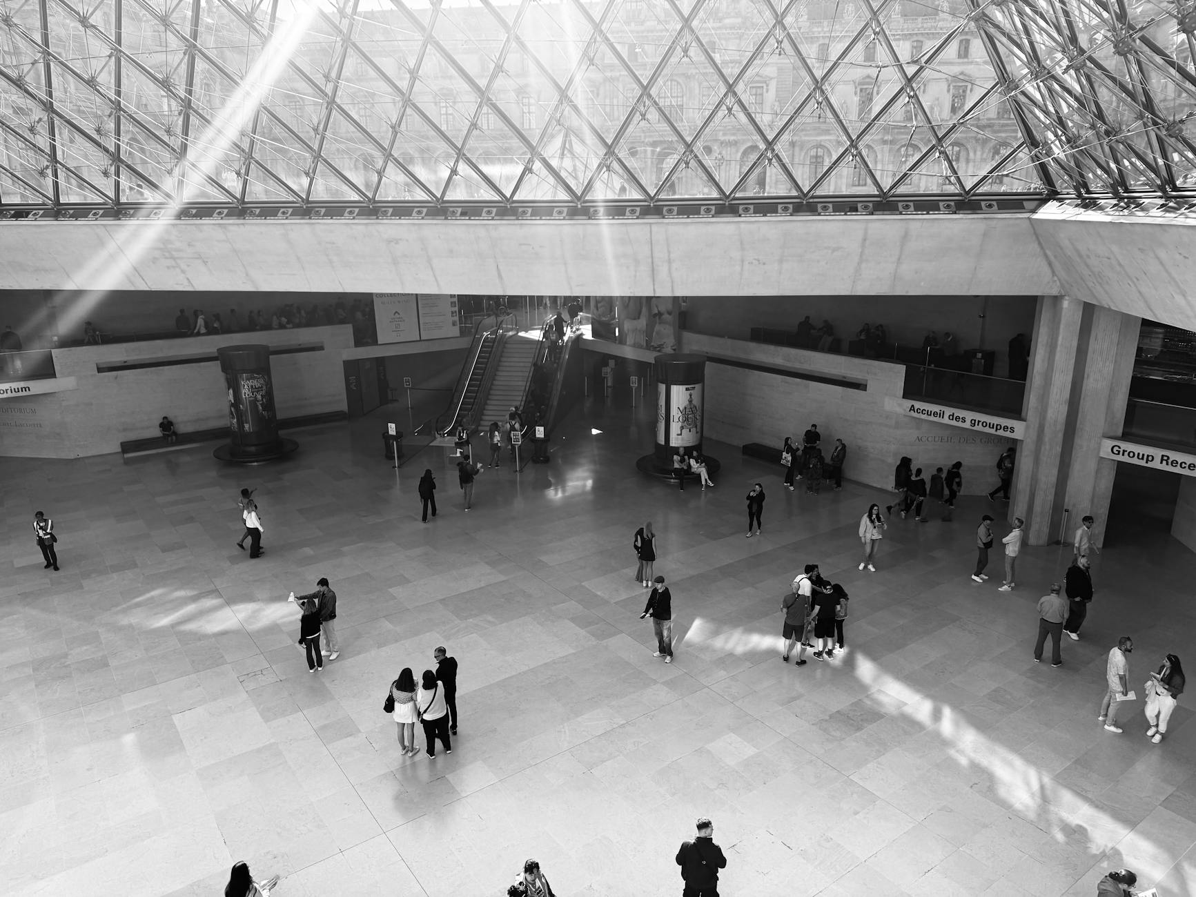 The glass atrium of the Louvre with visitors and streaming sunlight