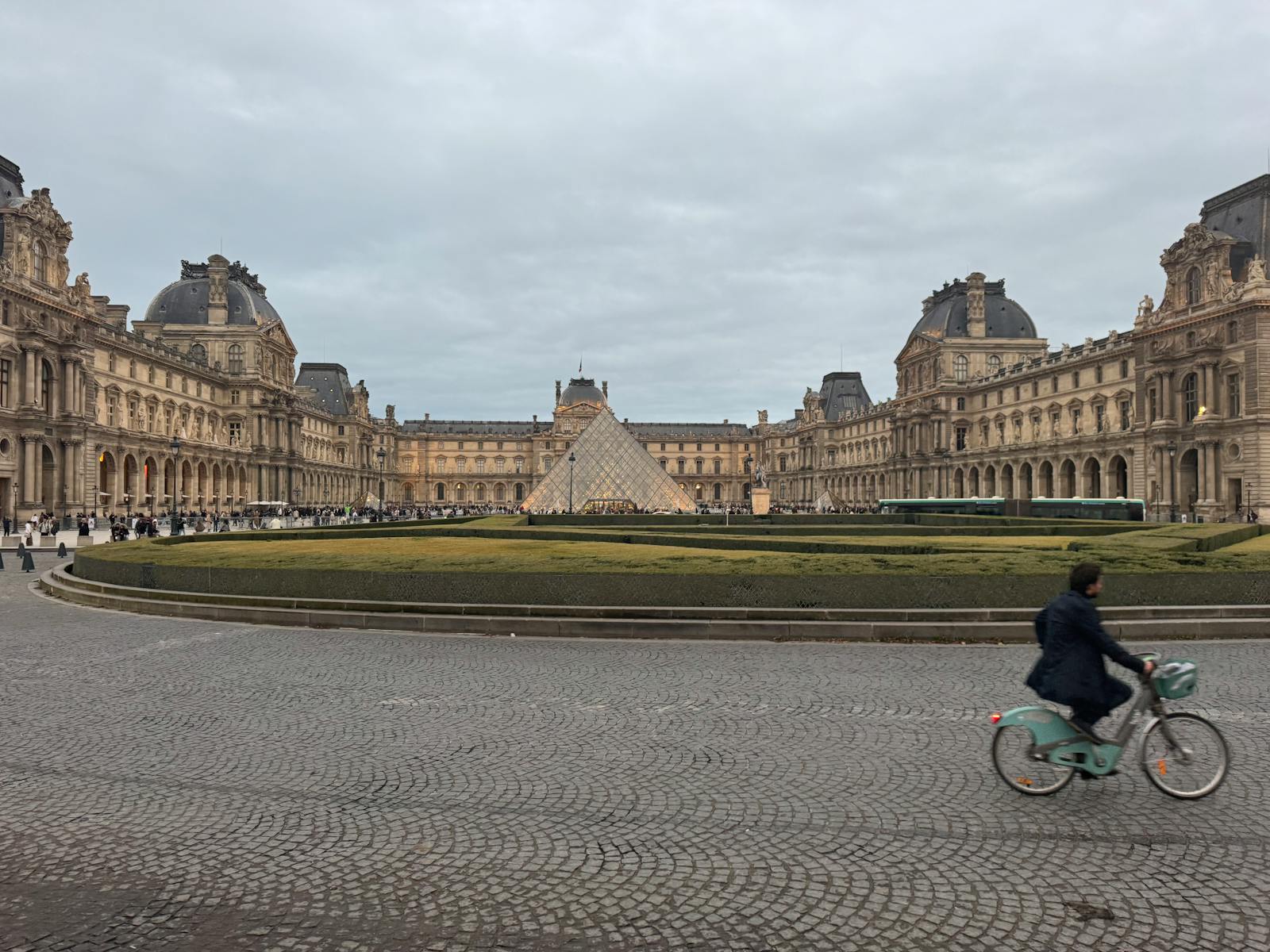 A cyclist rides past the Louvre Pyramid in Paris with classic French architecture in view
