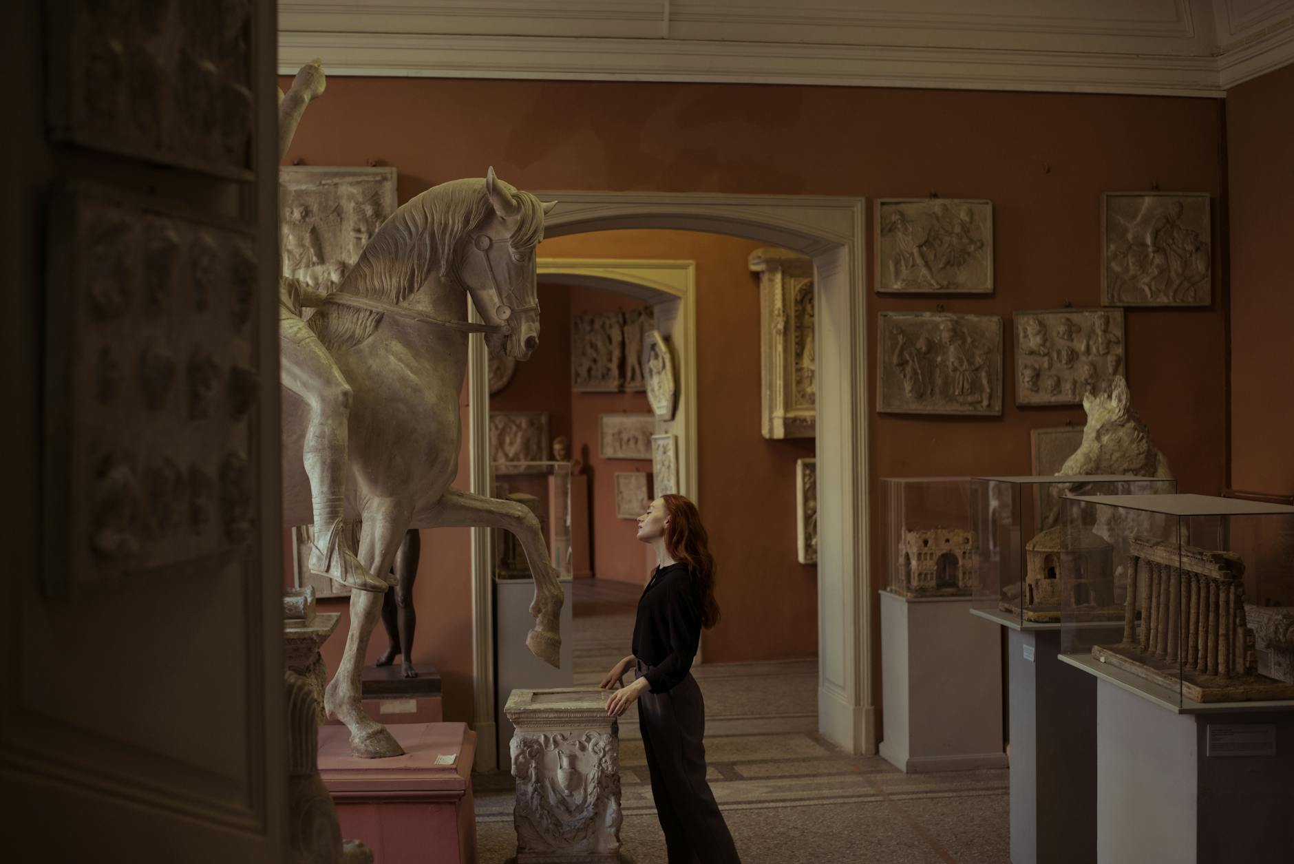 A woman admiring a horse sculpture in an art museum surrounded by classical artwork